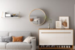 Een moderne woonkamer met een grijze bank, aardetinten kussens, een witte en houten kast, ingelijste kunst, de Spiegel 'Ria' 80x80 Oude eik (1c) spiegel, een vaas met een plant en een gouden plank met boeken op een lichtgrijze muur.