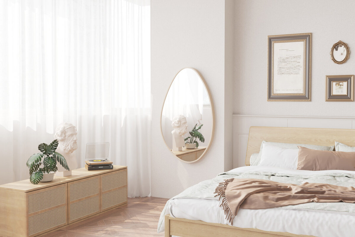 Een lichte, minimalistische slaapkamer heeft lichthouten meubels, een keurig opgemaakt bed, ingelijste kunst, een potplant en de ovale Spiegel 'Romi' 80x50 Oude eik spiegel boven een zijkast met een decoratieve buste door gordijnen.