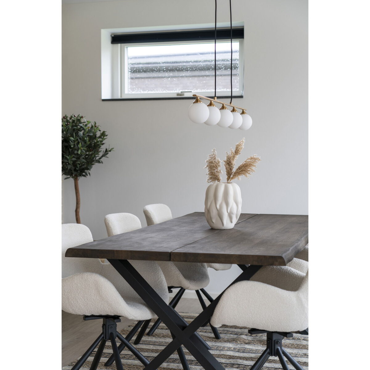 Een moderne eetkamer met een houten tafel, zes witte stoelen met kussens, een beige keramieken vaas (19,5x19,5x26 cm), gedroogde planten, een boom in pot in de hoek, een gestreept vloerkleed en een raam boven de tafel dat natuurlijk licht binnenlaat.
