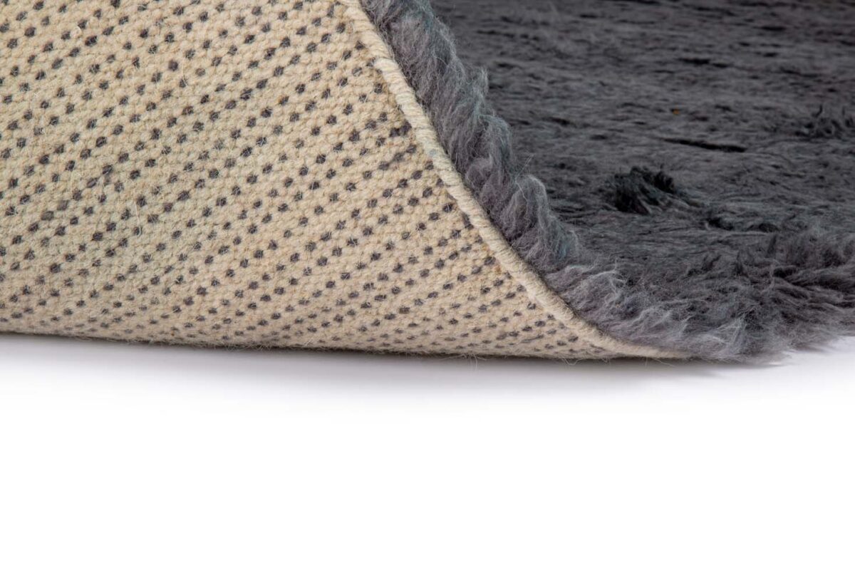 Een close-up van het Merano Charcoal 013 170x230 tapijt toont de donkergrijze, pluizige textuur, gedeeltelijk gevouwen om een beige onderkant met een gestippeld antislippatroon te onthullen.