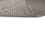Close-up van het Burano Dark Grey 618-613 200x300 vloerkleed, met een geweven structuurpatroon in lichtgrijs. Eén hoek is lichtjes gekruld om de dikte en het detail te onthullen tegen een witte achtergrond.