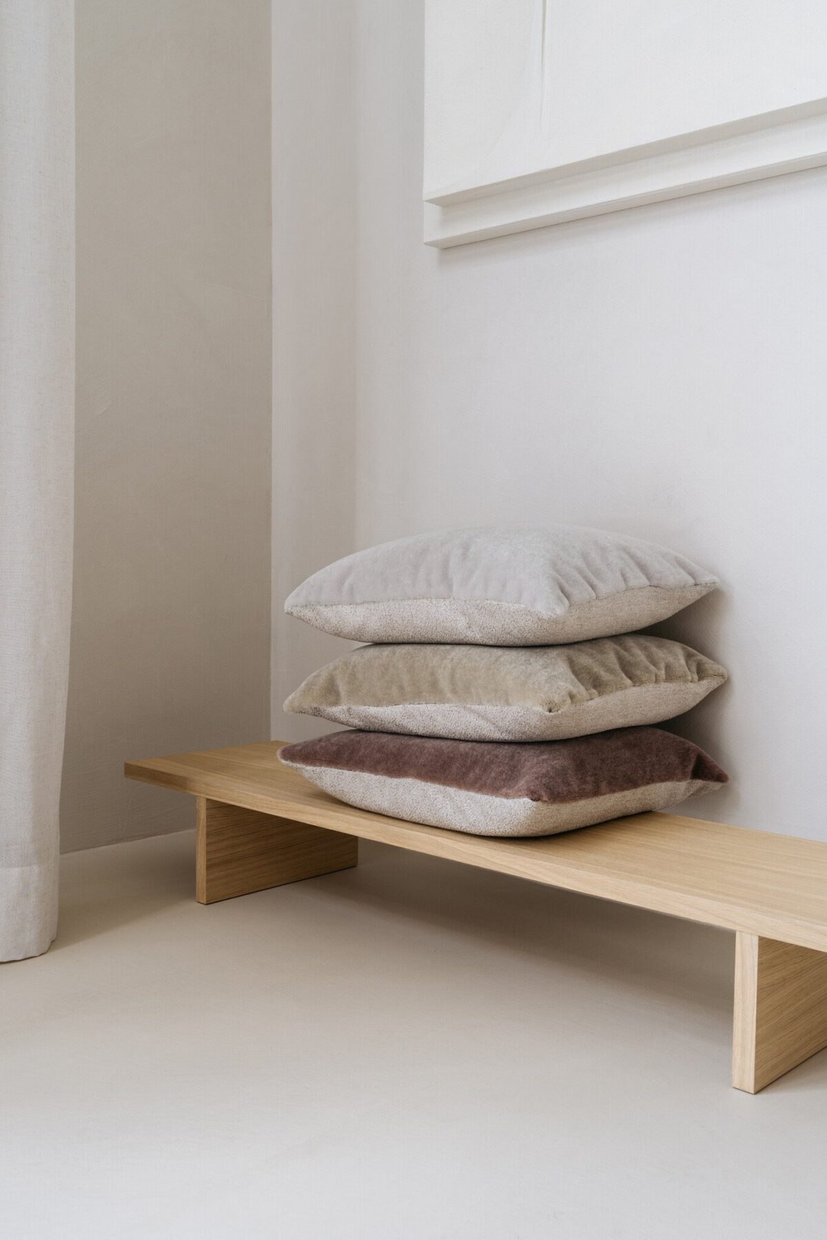 Een minimalistisch houten bankje toont een nette stapel van vier Mohair sierkussenhoes Amy grijsgroen 50x50 cm kussens tegen een lichte muur in een lichte, serene kamer.