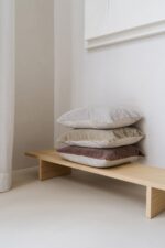 Een minimalistisch houten bankje toont een nette stapel van vier Mohair sierkussenhoes Amy grijsgroen 50x50 cm kussens tegen een lichte muur in een lichte, serene kamer.