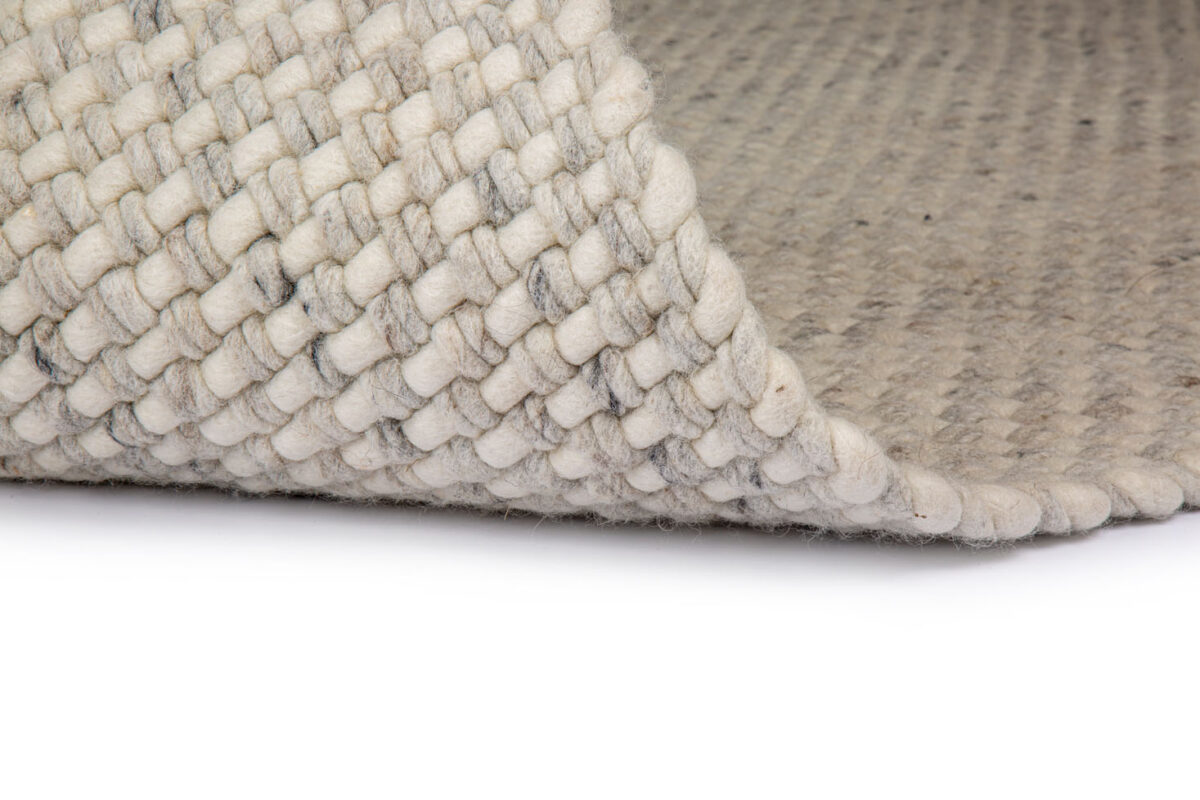Een close-up van het Burano White Grey 001-367 170x230 tapijt toont de lichte grof gebreide wollen textuur, met een hoek omgevouwen om de geweven onderkant en dikke garen details te onthullen.