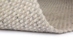 Een close-up van het Burano White Grey 001-367 170x230 tapijt toont de lichte grof gebreide wollen textuur, met een hoek omgevouwen om de geweven onderkant en dikke garen details te onthullen.