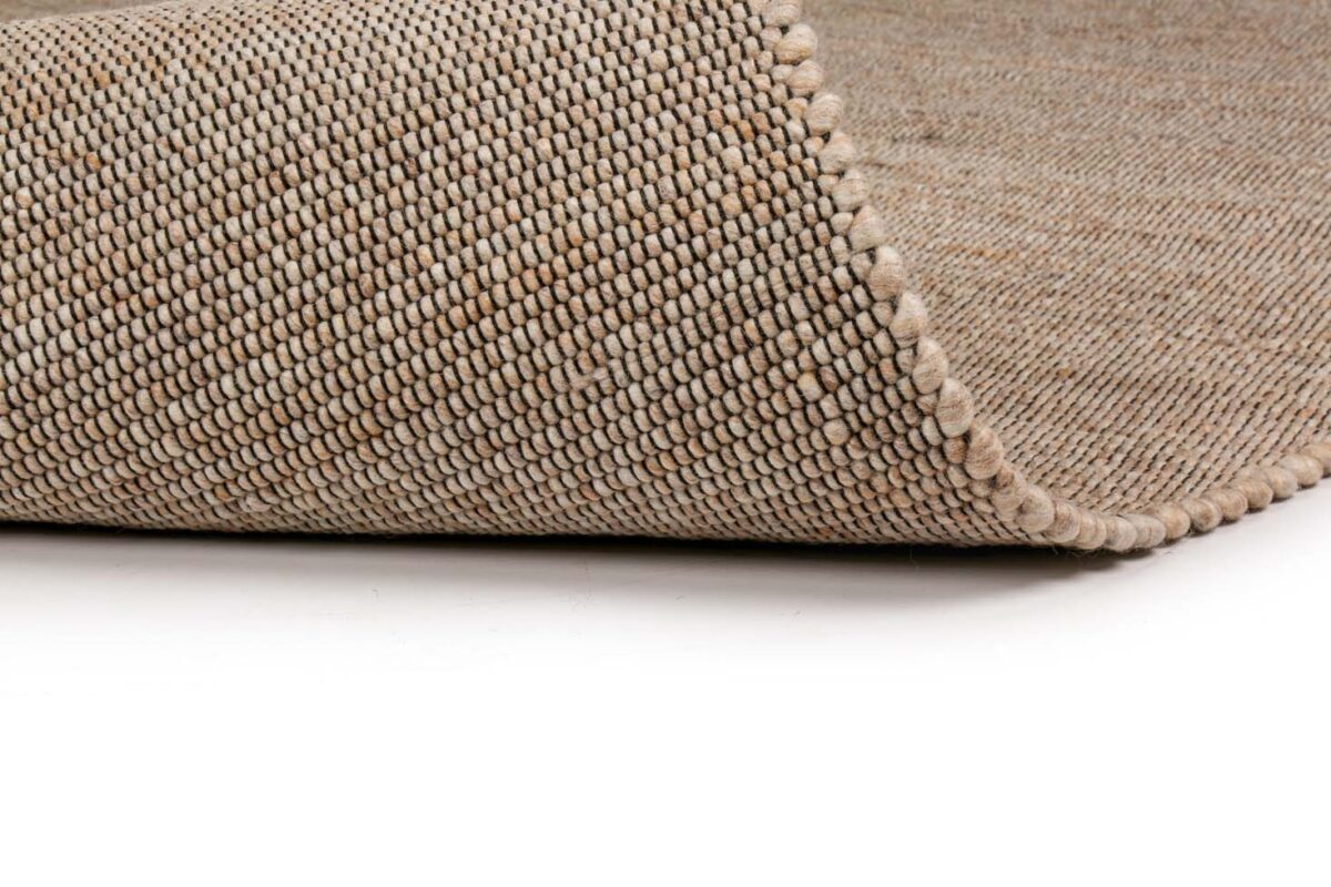 Close-up van het Bressano Beige 142 200x300 tapijt, met de nadruk op het geweven patroon en de netjes gestikte randen, met een hoek omhoog gekruld om de dikte van het materiaal te laten zien tegen een witte achtergrond.
