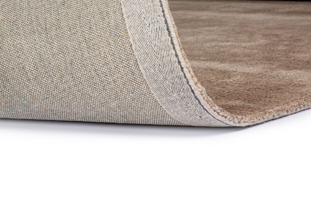 Close-up van de rand van het Rome Beige 02 200x300 tapijt met de hoek iets opgetild, waardoor het beige gestructureerde oppervlak, de onderkant en de dikte van het materiaal zichtbaar worden tegen een witte achtergrond.