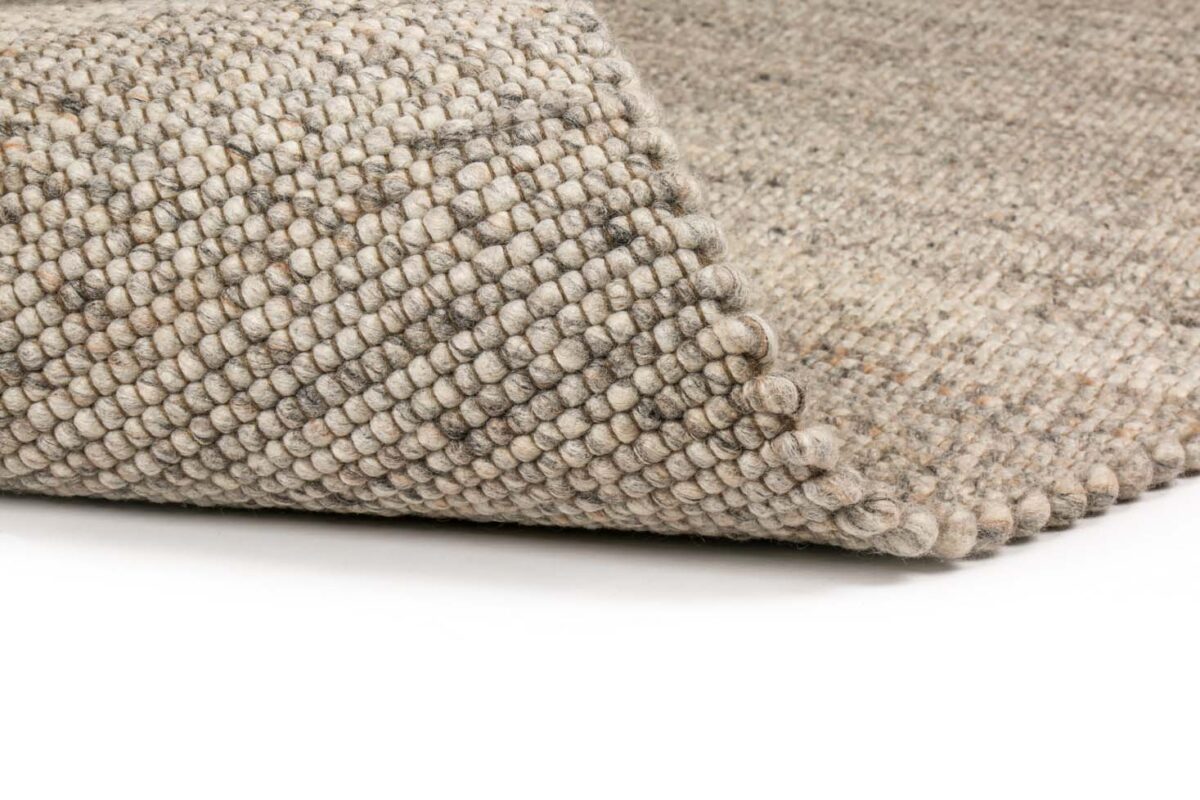 Close-up van de hoek van het Verona ST Natural Grey 834 170x230 tapijt, met de nadruk op het dikke, geluste, getextureerde weefsel van grijze wol. Het tapijt is licht opgetild om de dikte en de details te onthullen, getoond op een wit oppervlak.