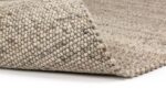 Close-up van de hoek van het Verona ST Natural Grey 834 170x230 tapijt, met de nadruk op het dikke, geluste, getextureerde weefsel van grijze wol. Het tapijt is licht opgetild om de dikte en de details te onthullen, getoond op een wit oppervlak.