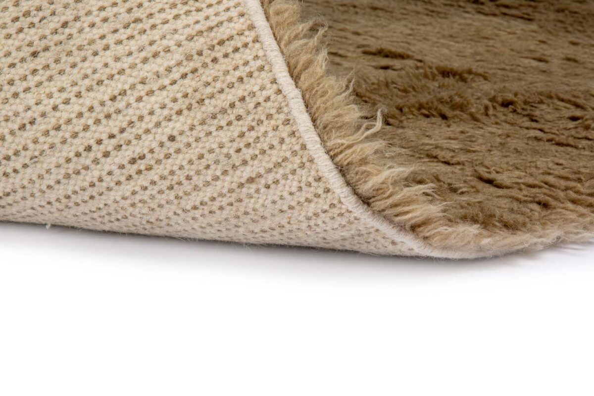 Close-up van het Merano Beige 002 200x300 tapijt met de hoek opgetild, waardoor een gestructureerde, slipvaste onderkant zichtbaar wordt tegen een witte achtergrond.