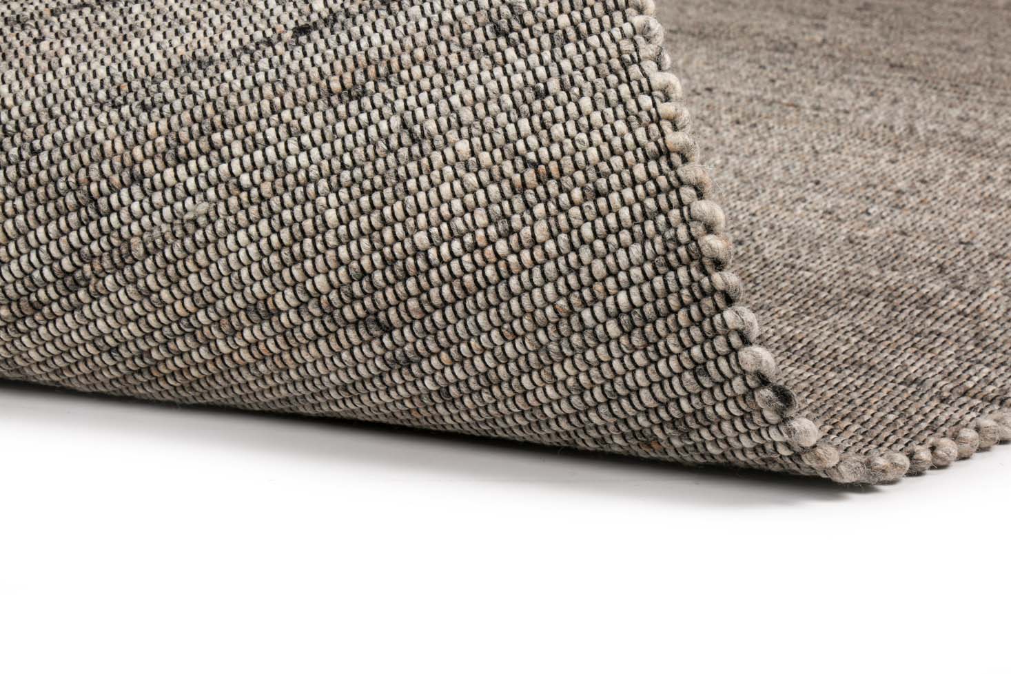 Een close-up van het tapijt Bressano Grey 834 170x230 in neutrale tinten, met een gevouwen hoek die het dikke, gedetailleerde geweven patroon tegen een witte achtergrond onthult.