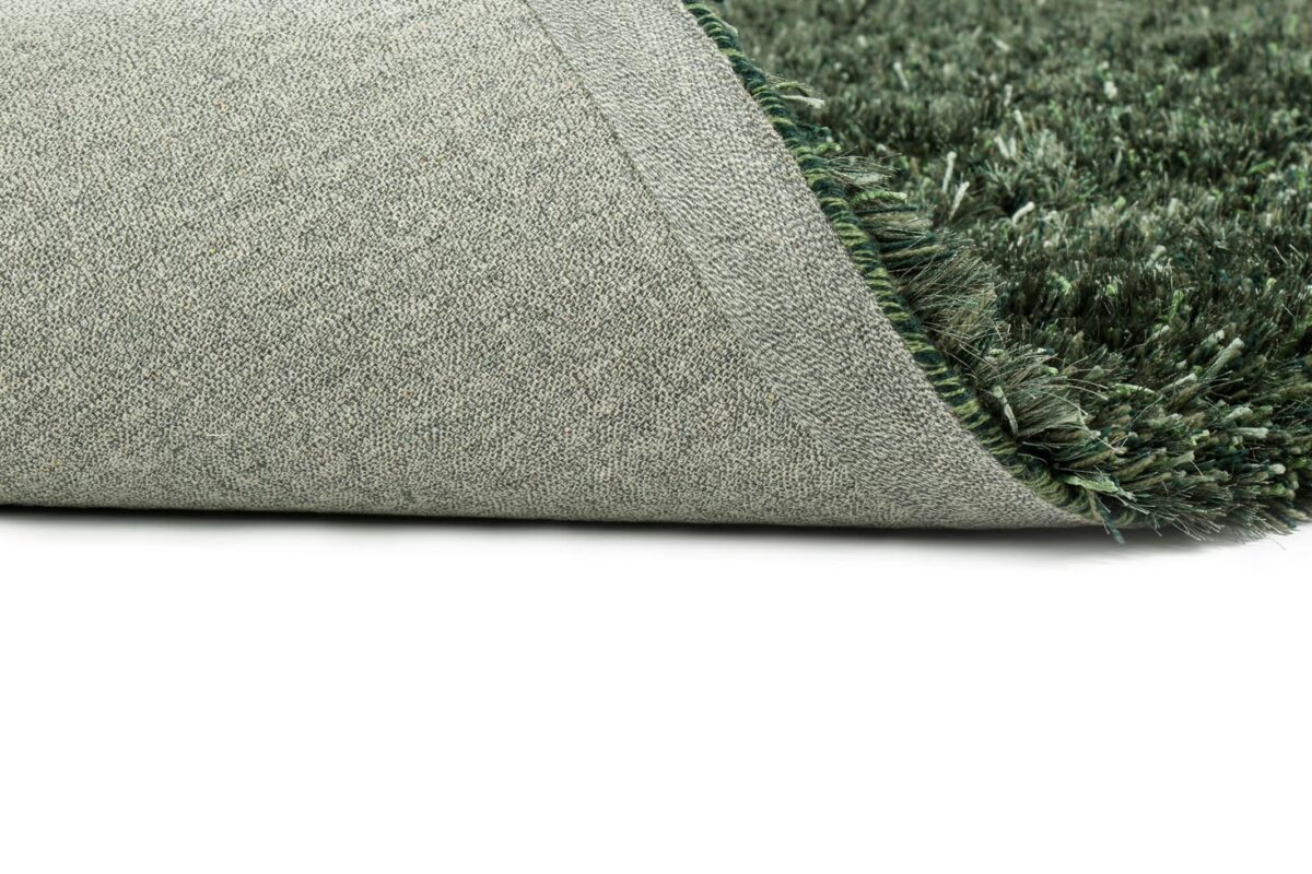 Een close-up van de hoek van het New Paulo Dark Green 442 170x230 vloerkleed omgedraaid, waardoor de dikke, zachte ruige vezels en de getextureerde onderkant zichtbaar worden tegen een witte achtergrond.