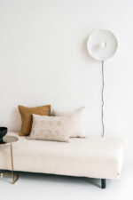 Minimalistisch interieur met een beige daybed met structuur, twee Sierkussenhoes Jane 60x40 cm neutrale kussens, een gouden bijzettafel en een witte wandlamp met zichtbaar snoer tegen een witte muur.