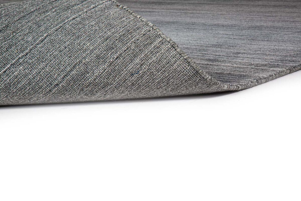 Een close-up van het Portofino Blue 01 200x300 tapijt toont het grijs gestructureerde oppervlak met een hoek omgevouwen om de onderkant te onthullen, tegen een witte achtergrond.