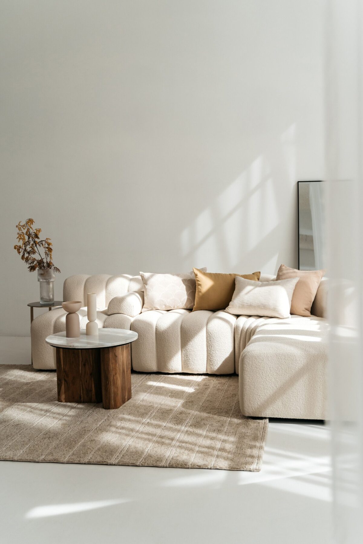 Minimalistische woonkamer met een beige sectionaal, Sierkussenhoes Hazel 60x40 cm kussens, een vloerkleed met structuur, twee ronde salontafels met houten onderstel, droogbloemen in een vaas en een grote spiegel tegen een witte muur die baadt in het zonlicht.