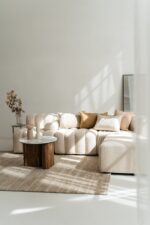 Minimalistische woonkamer met een beige sectionaal, Sierkussenhoes Hazel 60x40 cm kussens, een vloerkleed met structuur, twee ronde salontafels met houten onderstel, droogbloemen in een vaas en een grote spiegel tegen een witte muur die baadt in het zonlicht.