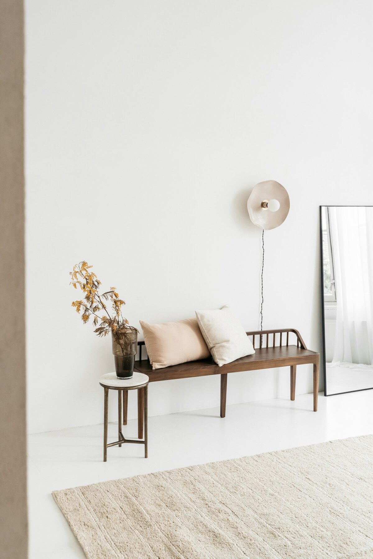 Minimalistische leefruimte met een houten bank, twee Sierkussenhoes Hazel 50x50 cm kussens, een kleine bijzettafel met droogbloemen, een ronde wandlamp, grote vloerspiegel en een beige vloerkleed op een witte vloer.