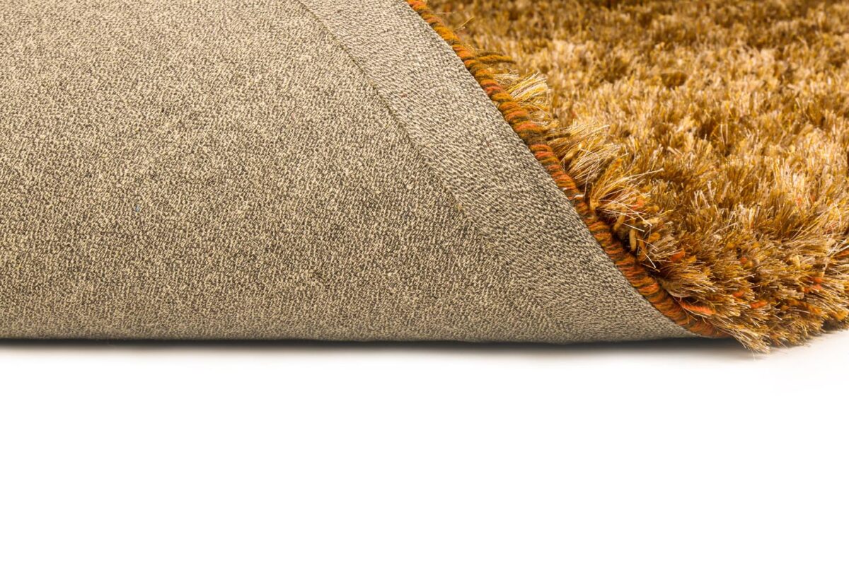 Close-up van het bruine shaggy vloerkleed New Paulo Gold 030 200x300 met een hoek omhoog, waardoor de gestructureerde achterkant en zachte, dikke pool zichtbaar worden tegen een witte achtergrond.