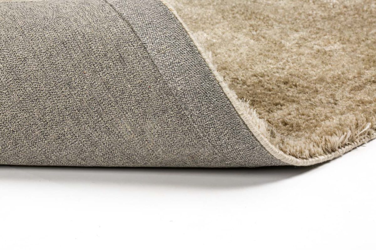 Een close-up van het beige Viterbo ST Silver 119 200x300 tapijt met een gestructureerd oppervlak en een gevouwen hoek die de grijs geweven onderkant onthult, tegen een witte achtergrond.