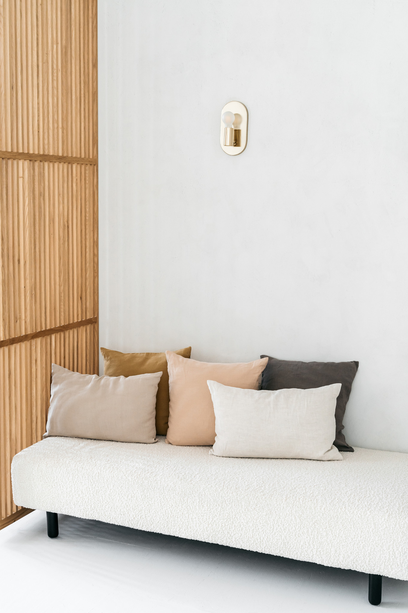 Een minimalistische woonruimte met een bank met witte structuur, vijf Sierkussenhoes Scotty Nude 60x40 cm kussens, een houten lattenbodem en een moderne wandlamp op een effen witte muur.