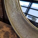 Een close-up van de Spiegel Rond Goud Medium toont de ronde, goudkleurige metalen lijst met een verweerde, gestructureerde afwerking. De spiegel weerspiegelt een raam en een blauwe lucht, terwijl op de voorgrond een deel van een houten oppervlak zichtbaar is.