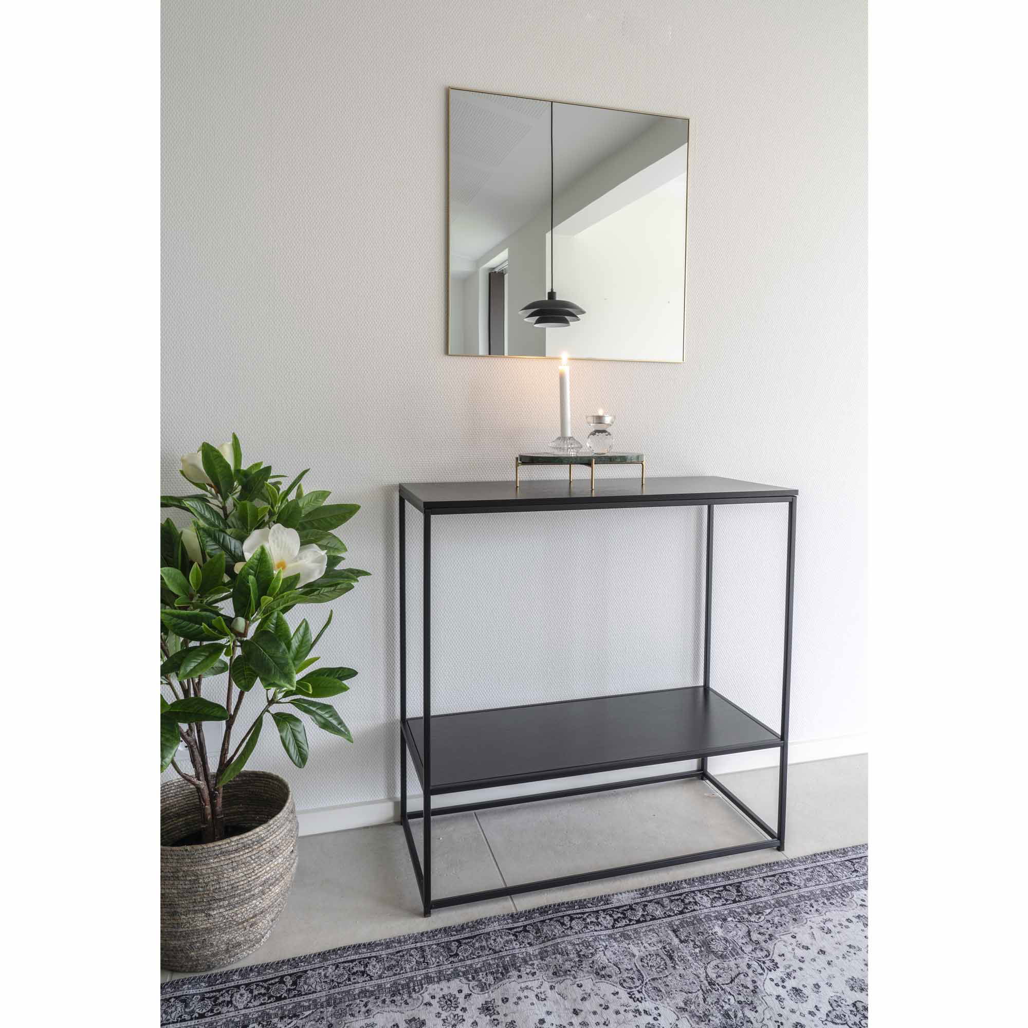Een minimalistische ingang heeft een zwart metalen consoletafel met glazen blad en plank, een plant in pot, de Jersey Mirror met een lijst in messinglook (60x60 cm) erboven en een kaars in een glazen houder op tafel.