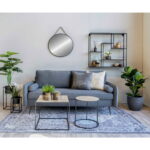 Moderne woonkamer met een grijze bank, decoratieve kussens, twee tafeltjes met potplanten, een vloerkleed met patroon, wandplanken, grote bladplanten in de hoek en de Trapani Mirror-zwart stalen frame en PU-band (Ø60 cm) aan de muur.