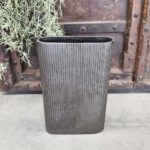 De Vaas Ovaal Loodkleurige 34, een hoge rechthoekige geribbelde grijze plantenbak, staat op beton met een rustieke houten muur en hangend groen op de achtergrond.