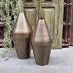 Twee Vaas Bronskleurig 49 vazen met een gehamerd metallic gouden afwerking staan naast elkaar op een betonnen vloer voor rustieke houten deuren met groene verf en hangend groen.