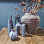 De Vaasje Organisch Blauw set van 4 bestaat uit gestructureerde vazen in moderne vormen en variërende grijstinten, gerangschikt op hout met gedroogde takken en zaaddozen in de grootste vaas, tegen een donkerblauw-grijze muur.