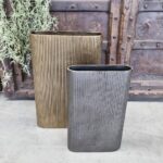 De Vaas Ovaal Bronskleurig 42, een rechthoekige geribbelde metalen vaas in brons, staat op beton voor een rustieke houten achtergrond met metalen accenten, met groen gebladerte dat aan de linkerkant hangt.