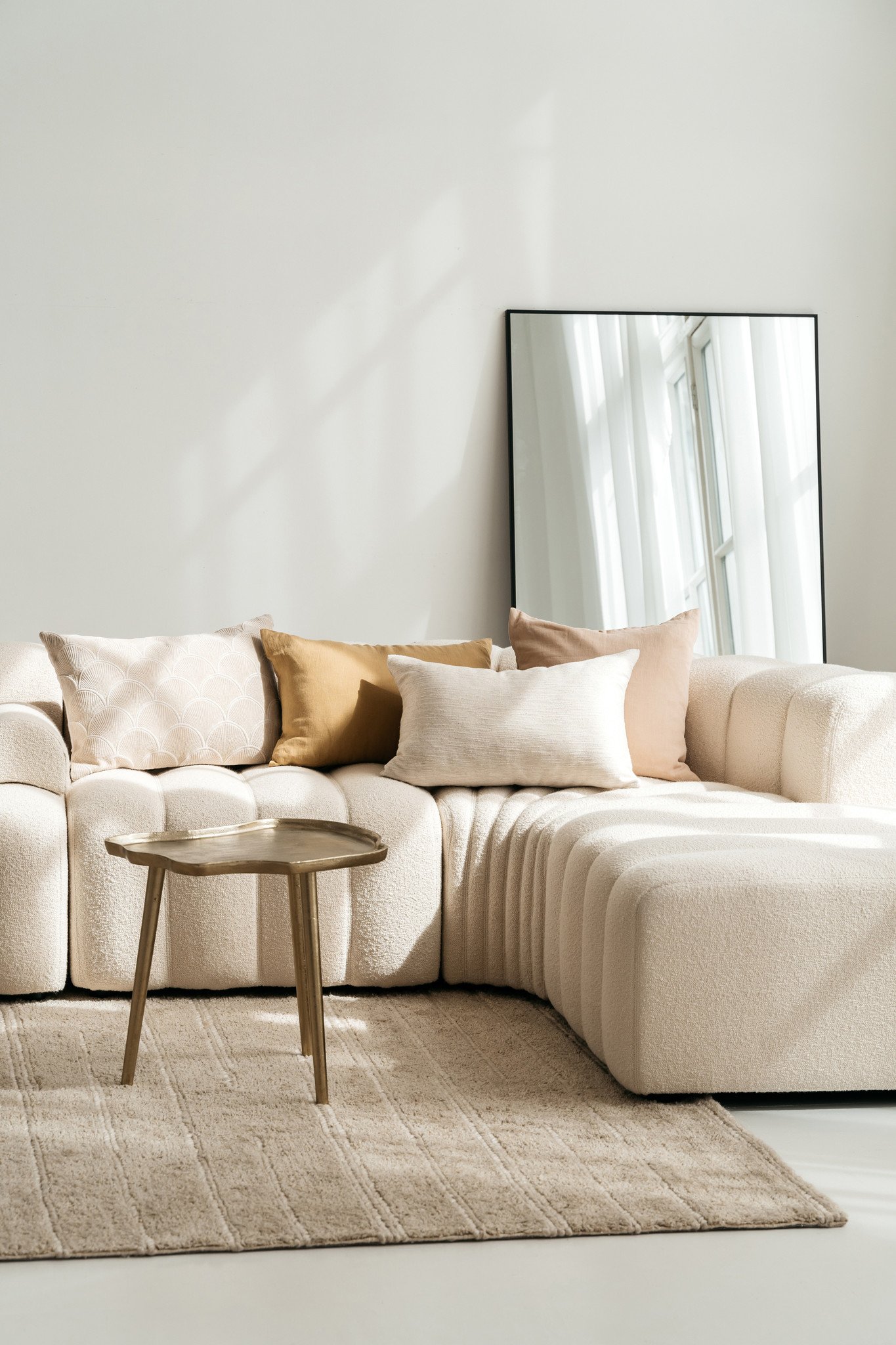 Een moderne woonkamer met een crèmekleurige sofa, neutrale kussens, een Sierkussenhoes Scotty Goudbruin 60x40 cm accentkussen, een beige vloerkleed, een kleine houten salontafel en een grote spiegel die het zonlicht van de ramen weerkaatst.