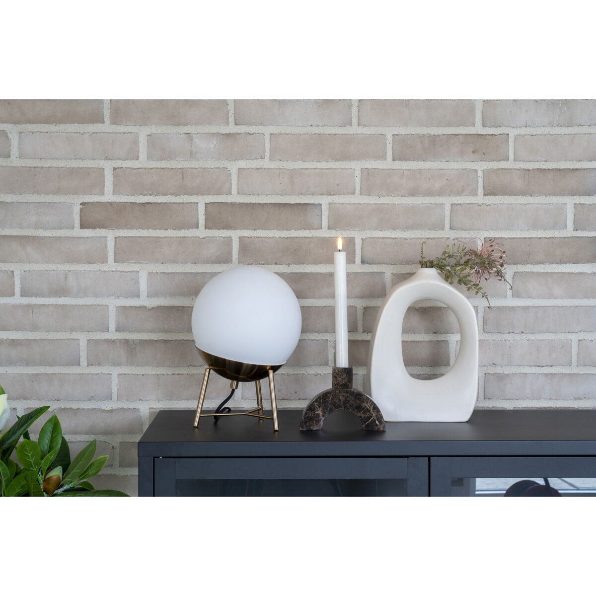 Een moderne lamp met een ronde witte kap, een brandende kaars in een donkere houder en de Vaas in organische vorm van wit keramiek 9x20x27,5 cm met bloemen staan op een donkere kast tegen een lichte bakstenen muur.