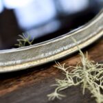 Een close-up van het Spiegel Organisch Goud, met de verweerde metalen rand rustend op hout, met delicate pluizige plantenstengels op de voorgrond.