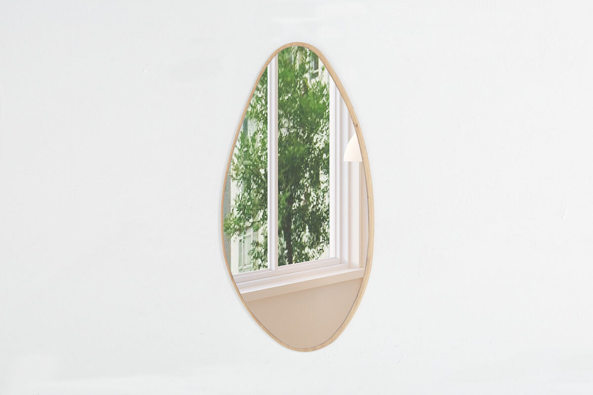 De Spiegel 'Rana' 80x50 Oude eik (1c), een ovale wandspiegel met een licht eikenhouten lijst, is gemonteerd op een effen witte muur en weerspiegelt een raam en groen gebladerte buiten.