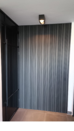 Moderne ingang met Rosenwood Akupanel Akoestische Wandpanelen Lattenwand Tripple Akupanelen 270 x 60 cm Zwarte eiken aan de muur, een zwart omlijste glazen deur die iets open staat aan de linkerkant, bruine vloertegels en een zwarte plafondlamp.