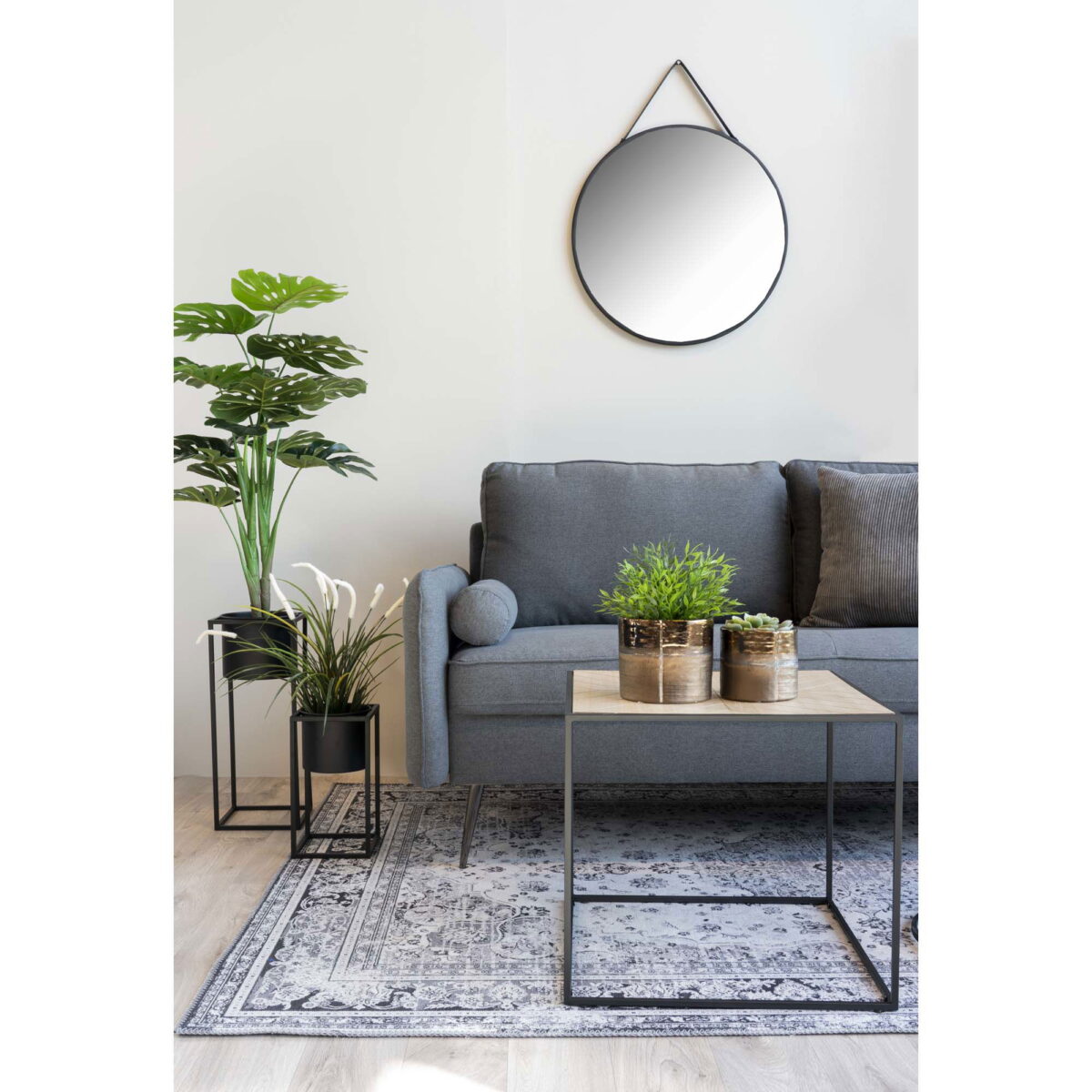 Een moderne woonkamer met een grijze bank, houten salontafel met twee potplanten, vloerkleed met patroon, vloerplant en de Trapani spiegel (zwart stalen frame, PU band, Ø60 cm) boven de bank.