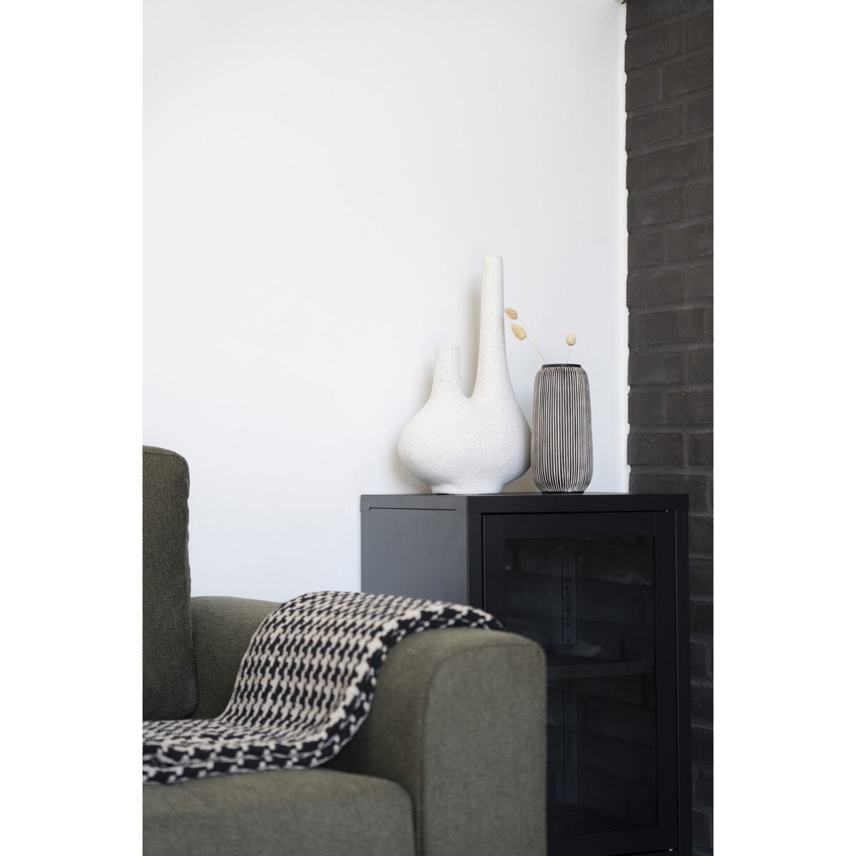 Een moderne woonkamerhoek heeft een groene fauteuil, een zwart-witte houndstooth deken en een zwarte kast met daarop de Vaas in keramiek, zwart/wit, rond, Ø9,5x20 cm, plus andere decoratieve vazen tegen een witte en donkere bakstenen muur.