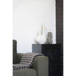 Een moderne woonkamerhoek heeft een groene fauteuil, een zwart-witte houndstooth deken en een zwarte kast met daarop de Vaas in keramiek, zwart/wit, rond, Ø9,5x20 cm, plus andere decoratieve vazen tegen een witte en donkere bakstenen muur.