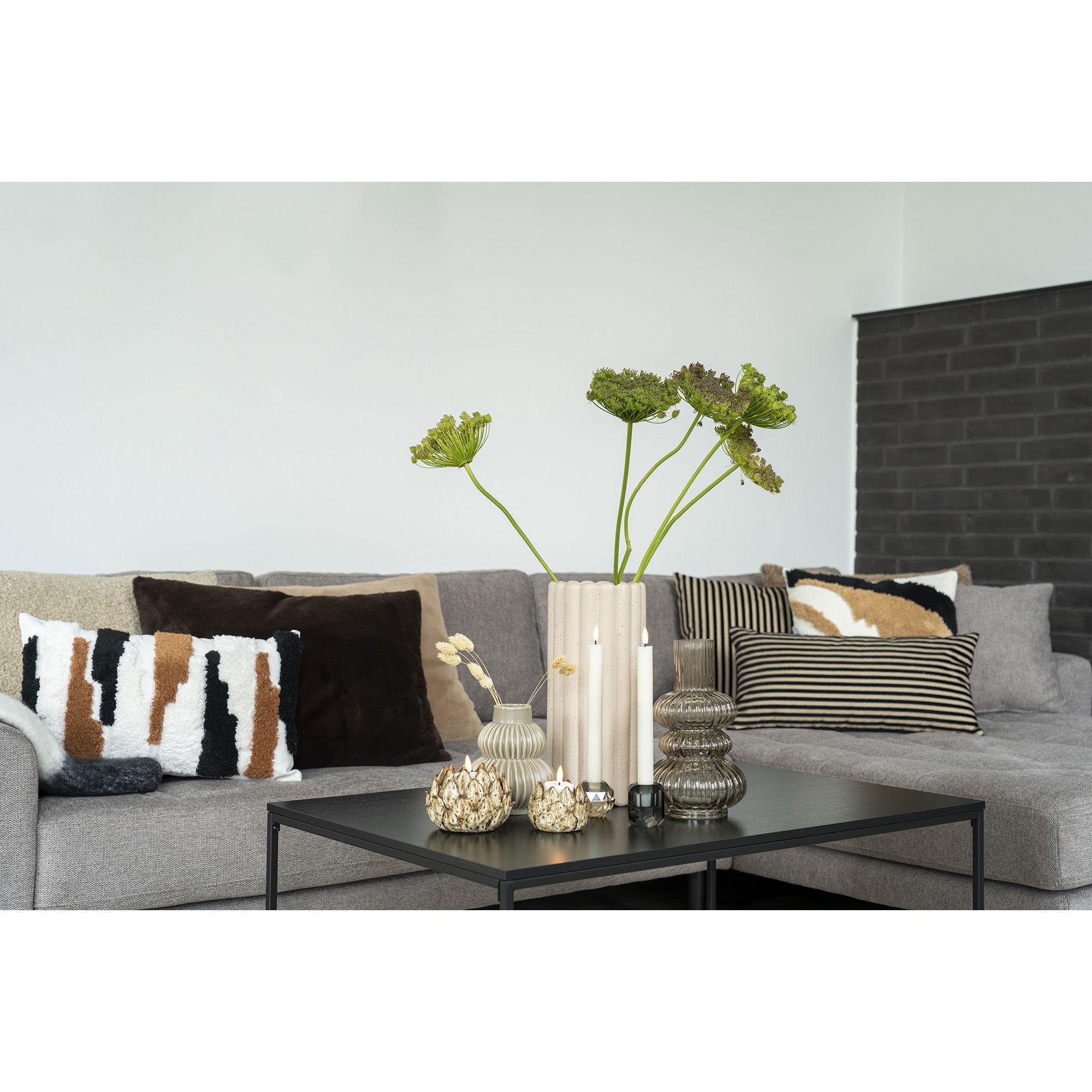 Een moderne woonkamer met een grijze bank met gedessineerde en effen kussens, een zwarte salontafel met vazen en kaarsen, groene planten in een Vaas - Vaas in gerookt mondgeblazen glas Ø15x25 cm, en een donkere bakstenen muur op de achtergrond.
