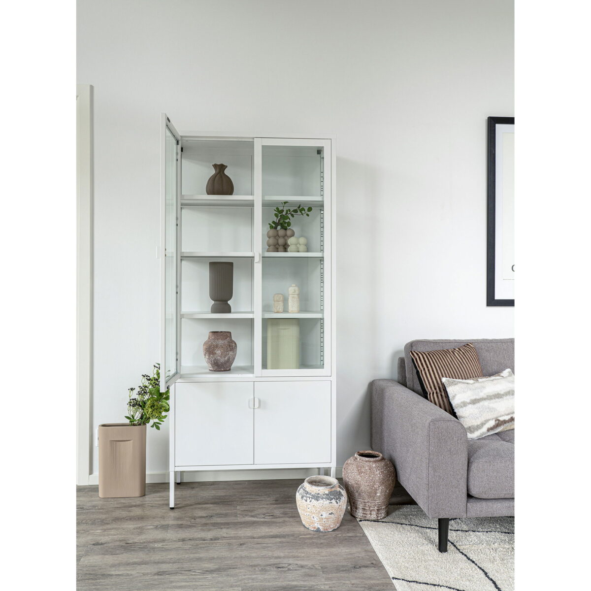Een witte vitrinekast met glazen deuren bevat decoratieve vazen, waaronder een vaas van grijs keramiek (Ø12x25,5 cm), en een plant. De kast staat naast een grijze bank met kussens met een patroon; andere vazen en planten staan op de houten vloer in de lichte kamer.