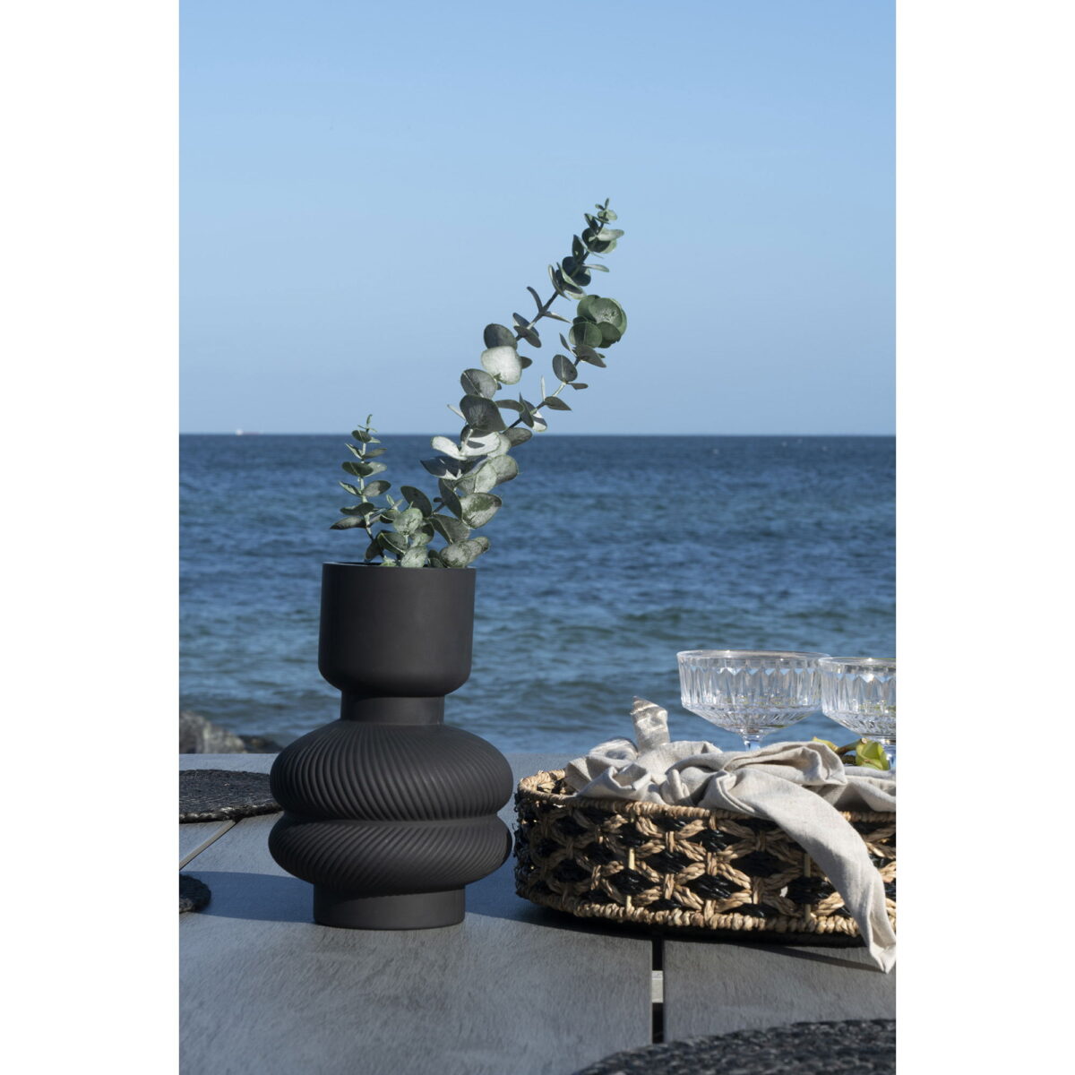 Een vaas van zwart glas Ø15x22 cm houdt groene eucalyptustakken vast op een tafel aan zee, vergezeld van gevlochten manden en helder glaswerk, met de oceaan en blauwe lucht zichtbaar op de achtergrond.