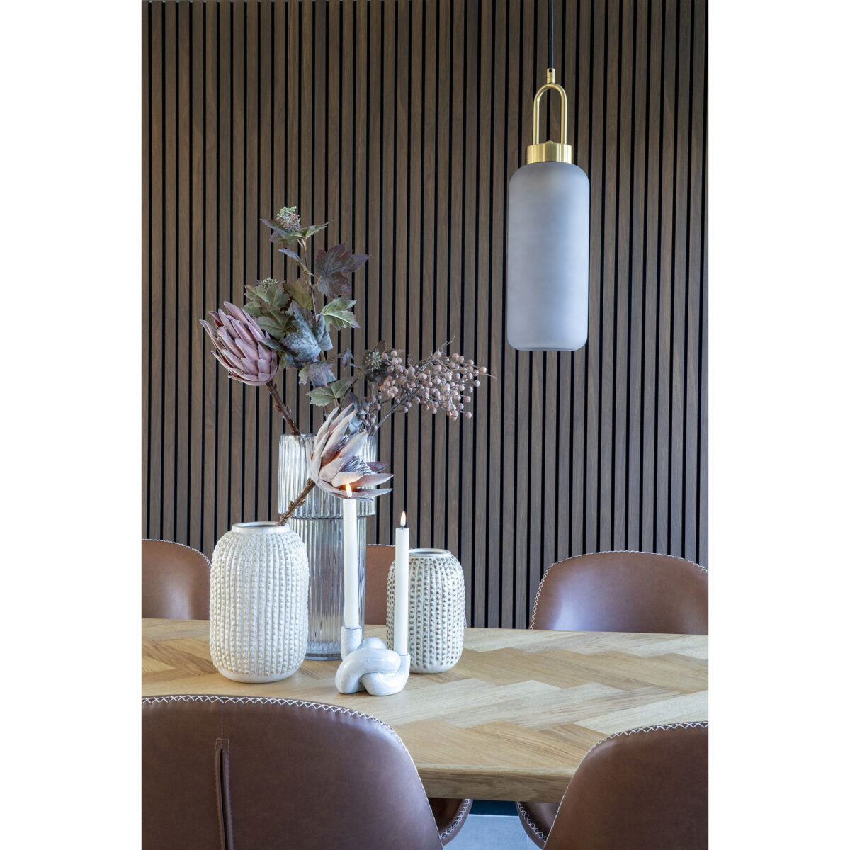 Een moderne eettafel met bruine leren stoelen, een ronde keramische vaas met beige patroon (Ø16x25,5 cm) met daarin gedroogde bloemen, een sculpturale kaars en een hoge matte hanglamp. De achtergrond bestaat uit verticale houten latten.