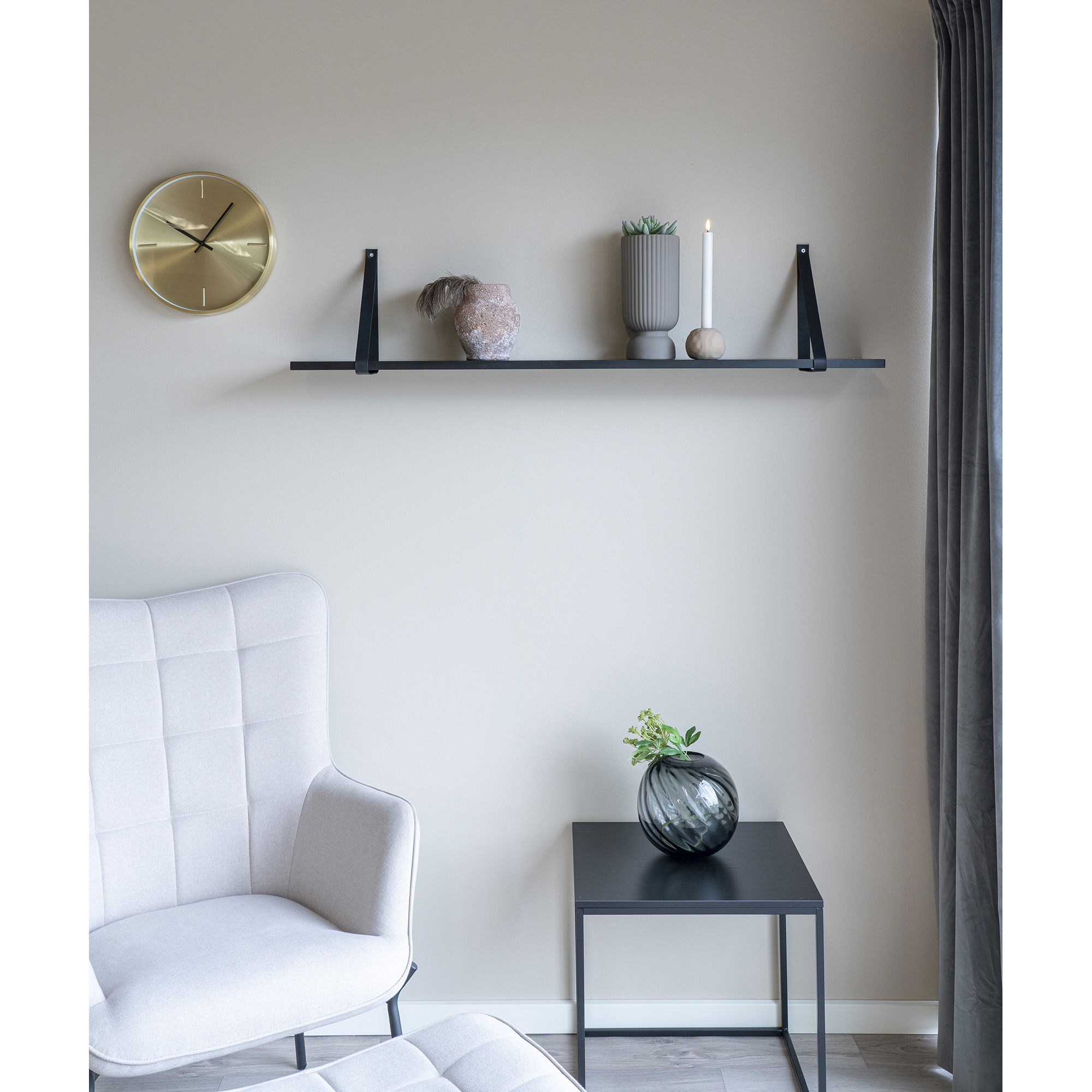 Een moderne minimalistische kamer heeft een beige fauteuil, een kleine zwarte bijzettafel met de Vaas in mondgeblazen glas, grijs, rond, Ø20x19 cm, een wandplank met decoratieve items en een gouden klok op een lichtgrijze muur bij een raam met grijze gordijnen.