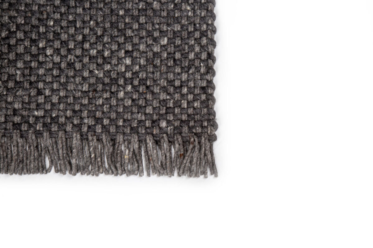Een close-up van de donkergrijze geweven hoek van het Burano Anthracite 614-604 170x230 vloerkleed toont de textuur en franjes tegen een effen witte achtergrond.