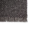 Een close-up van de donkergrijze geweven hoek van het Burano Anthracite 614-604 170x230 vloerkleed toont de textuur en franjes tegen een effen witte achtergrond.