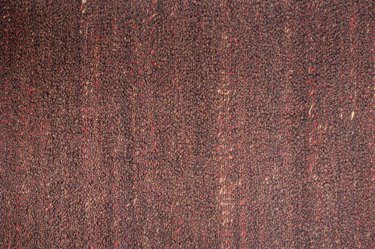Modena Bruin Antraciet 522 200x300 heeft een gestructureerd oppervlak met een dichte vlek van kleine, donkerbruine zaden, geaccentueerd door af en toe rode en bruine zaden voor een natuurlijke, organische uitstraling.