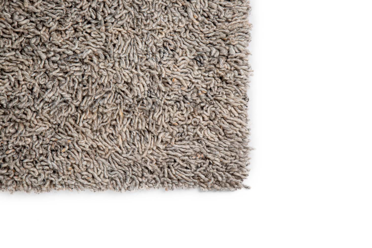 Close-up van de hoek van het Berbero Lungo Natural Grey 834 200x300 vloerkleed, met de dikke shaggy vezels tegen een witte achtergrond.