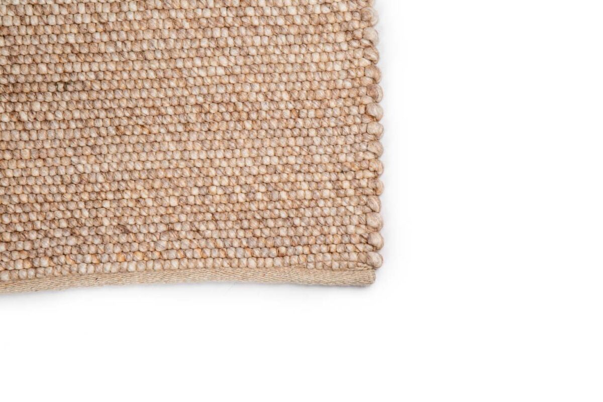 Close-up van de hoek van het Verona ST Beige 142 200x300 tapijt, met een grof geweven structuurpatroon op een beige achtergrond, tegen een witte achtergrond.