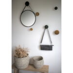 Een Trapani spiegel (zwart stalen frame, PU band, Ø38cm) en een kleine zwarte tas hangen aan een witte muur met ronde haken. Beneden staan twee gevlochten manden - een grote met gedroogde planten, een kleine - op een houten bank.